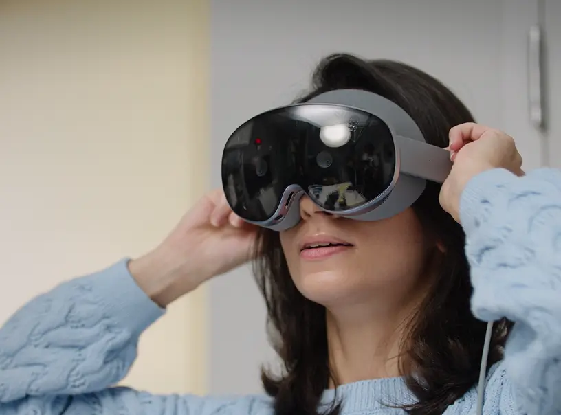 A woman with long dark hair wearing a light blue knit sweater, adjusting a grey Samsung Galaxy XR headset over her eyes with both hands.